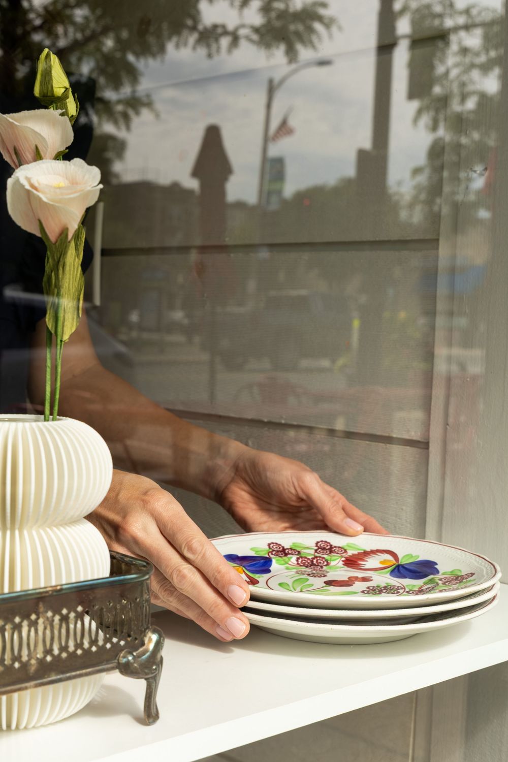 Hands arranging decorative floral plates on a white shelf beside a vase with white roses near a window.Swell House La Grange