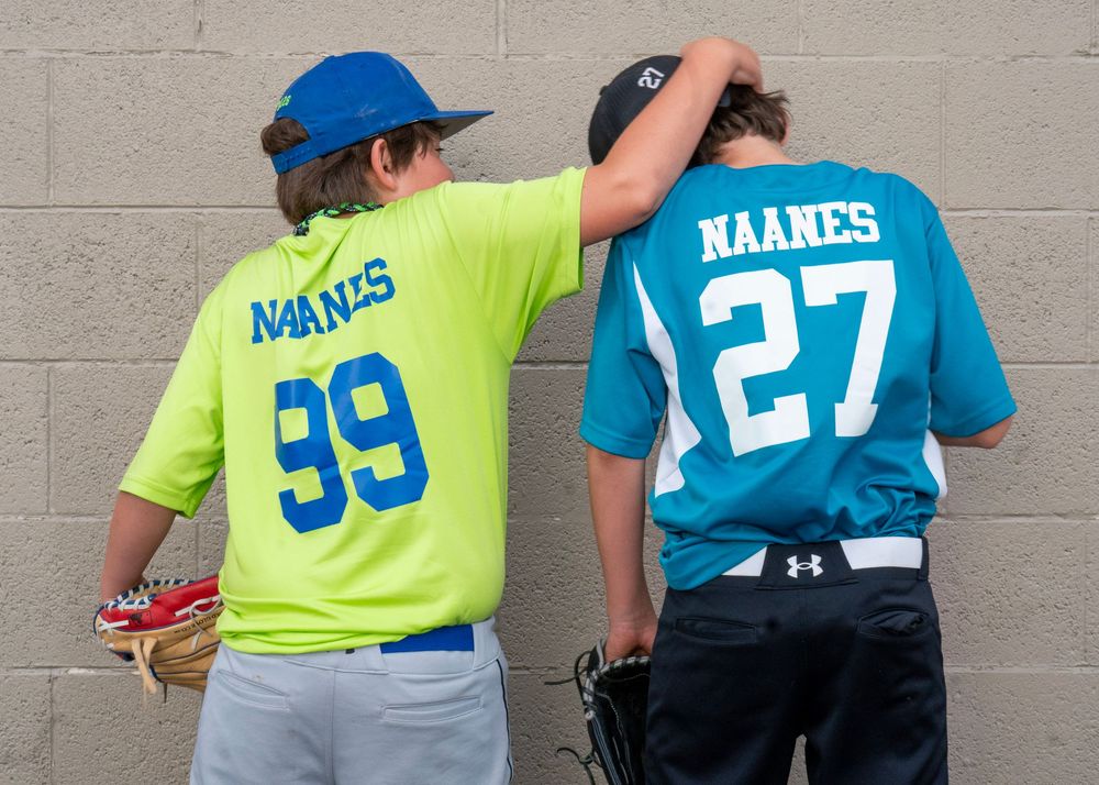 Two youth baseball players wearing Naanes jerseys numbered 99 and 27 stand against a wall.