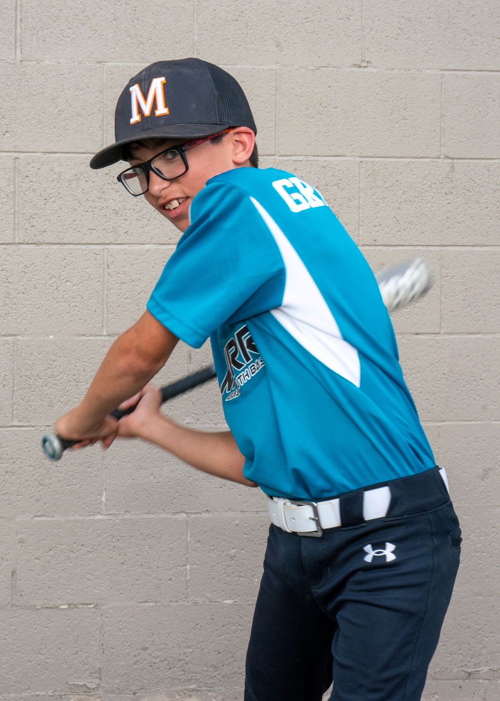 Young baseball player in teal uniform swinging a bat wearing glasses and an M cap.