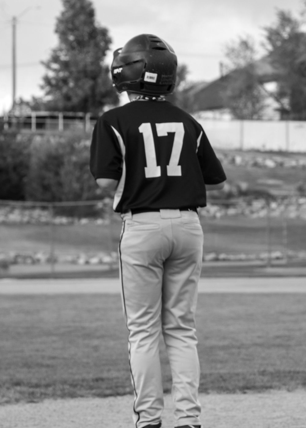 Young baseball player wearing number 17 jersey and batting helmet stands on field viewed from behind.