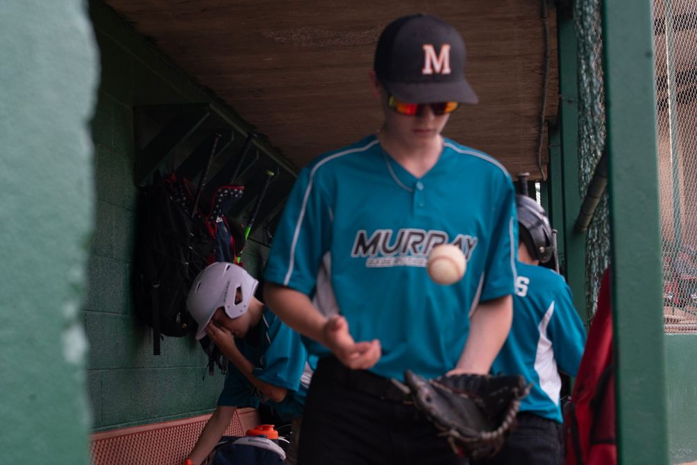A baseball player in a teal Murray jersey tosses a ball in the dugout.