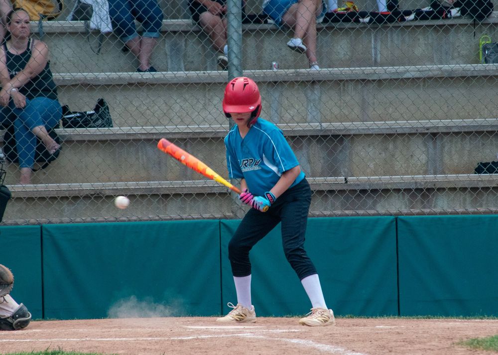 Young baseball batter in teal uniform and red helmet swinging at pitch during youth league game.