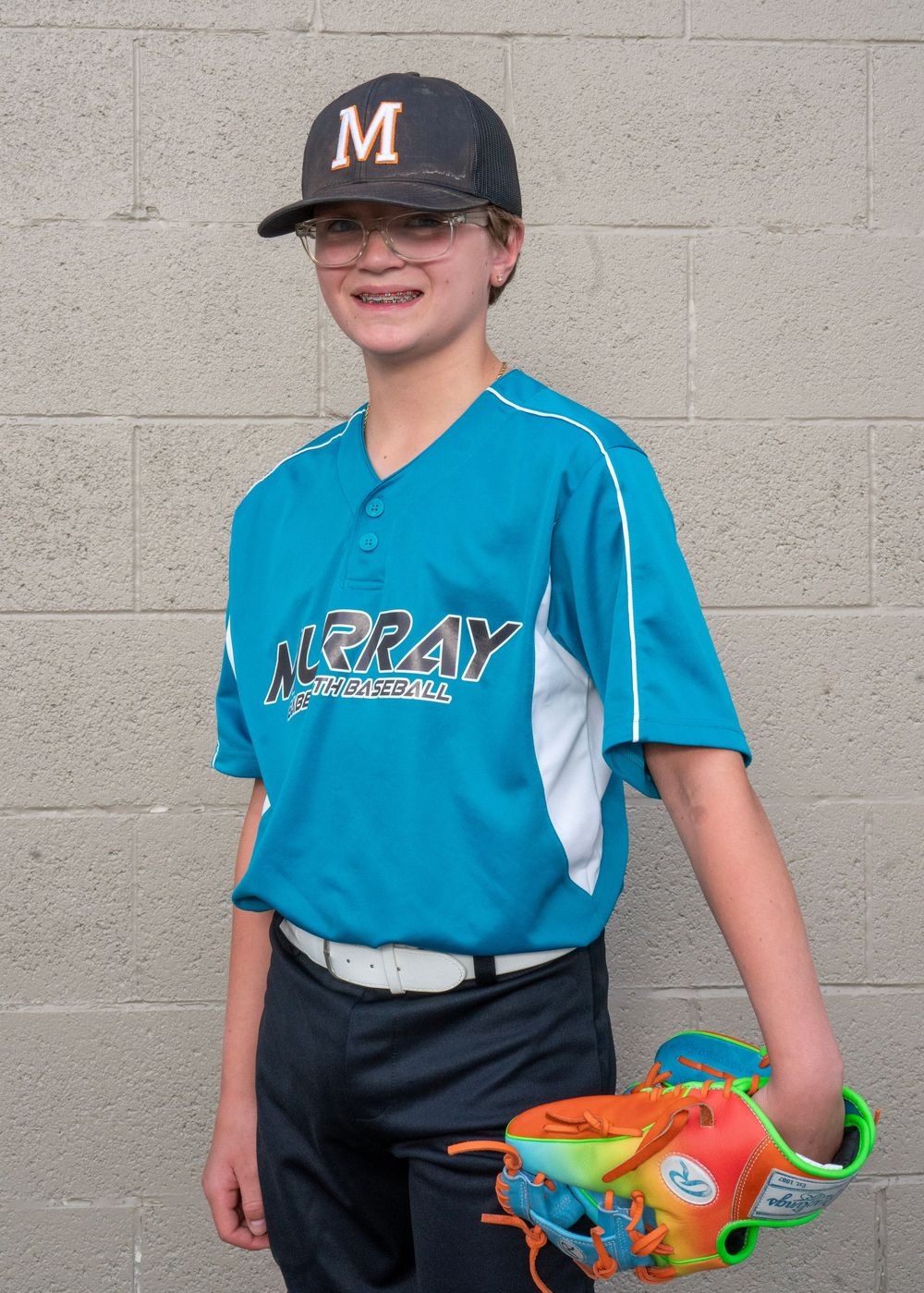 Young baseball player in teal Murray Baseball jersey and cap, smiling while holding a colorful glove against a gray wall.