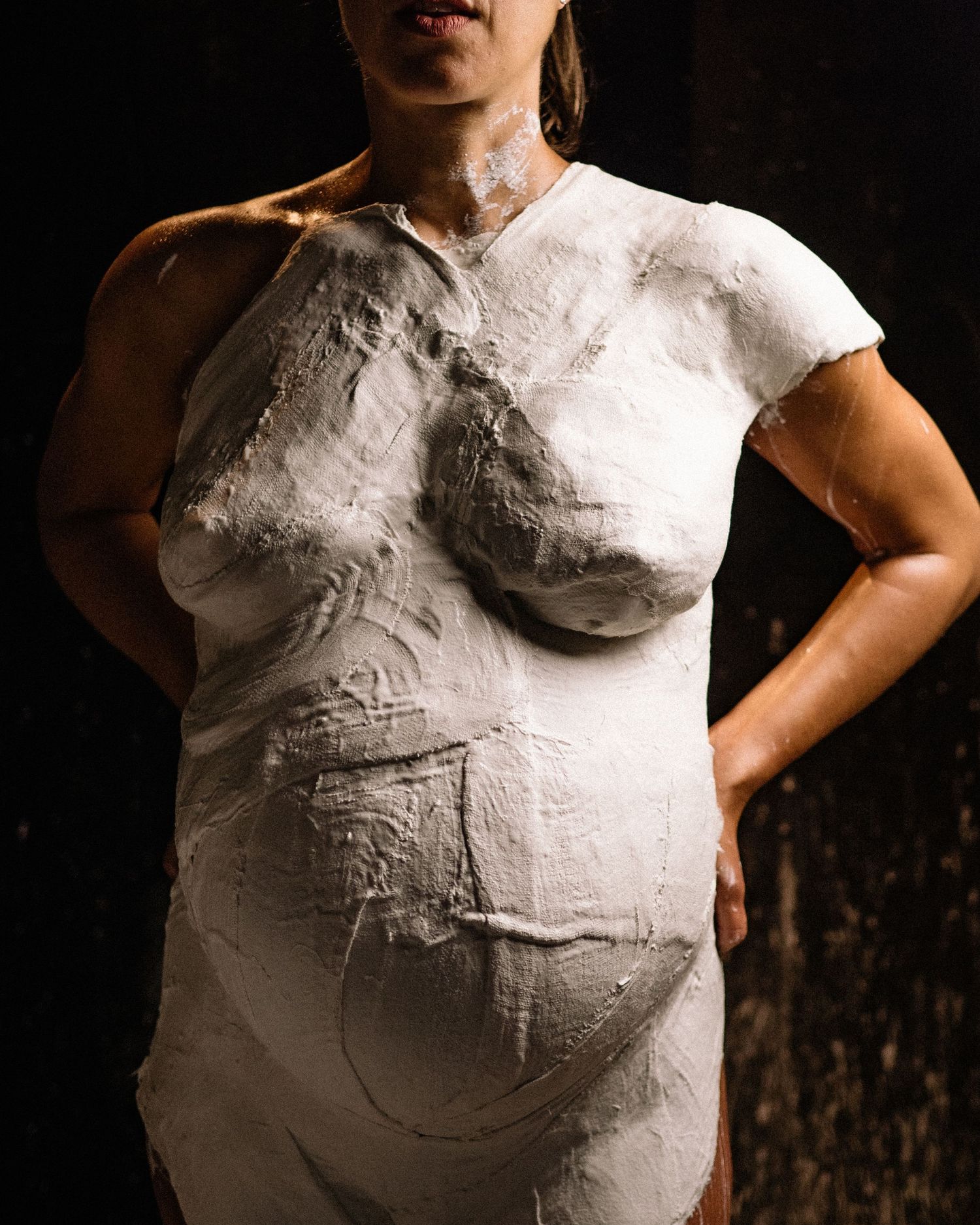 Pregnant woman torso covered in white plaster cast, artistic maternity body mold sculpture on dark background.