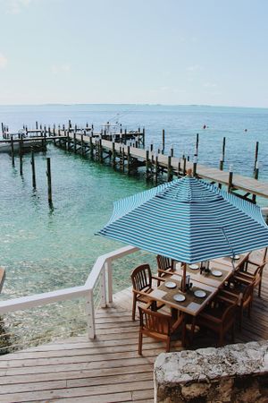 Wooden pier extends over turquoise water with striped umbrella dining area in foreground.