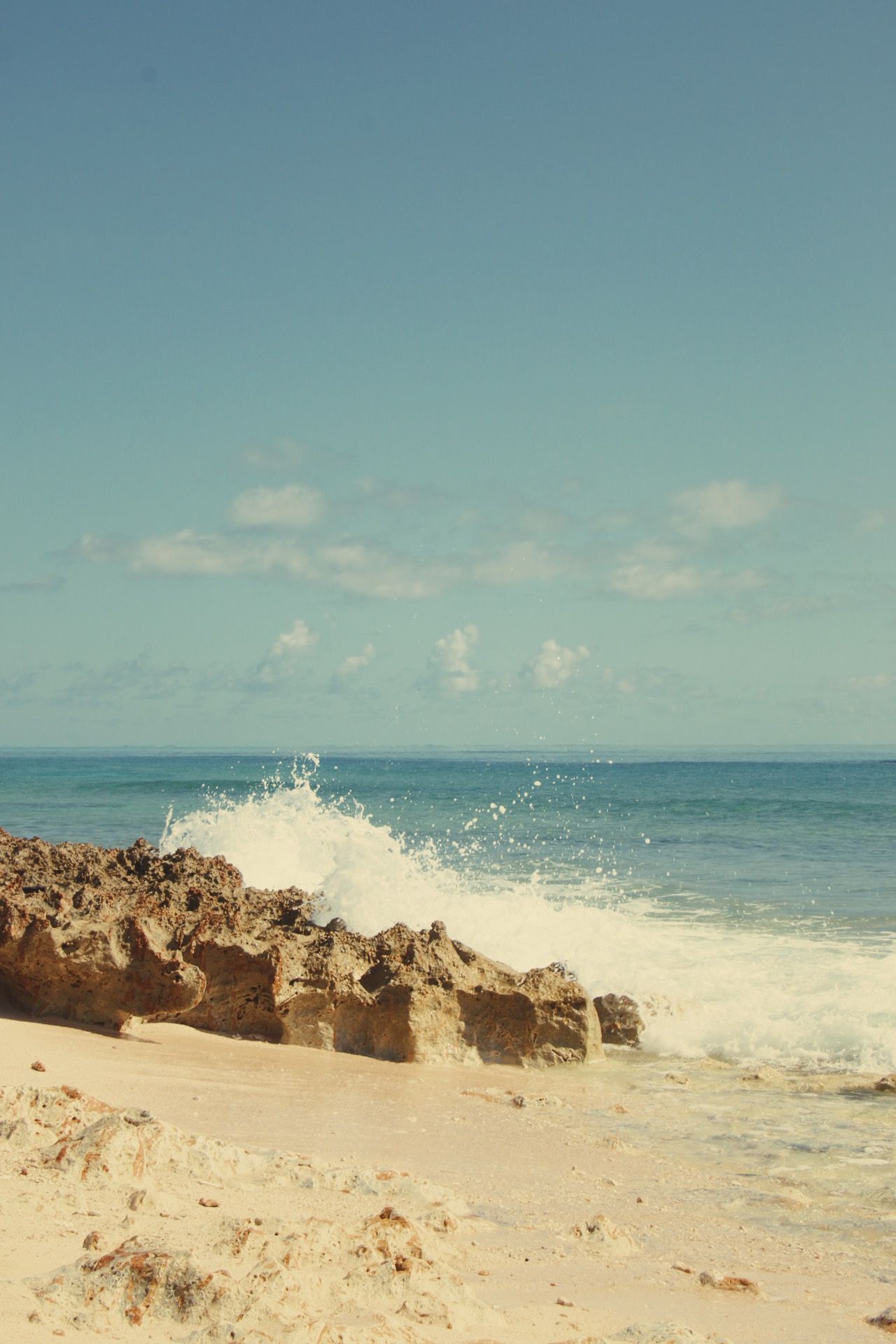 Ocean waves crash against rocky shore creating dramatic splash under vintage-toned sky.