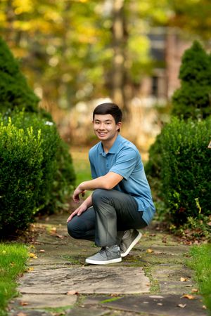 Smiling young man crouching on stone garden path surrounded by lush green shrubs.