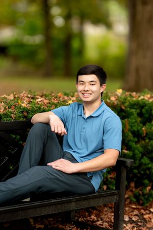 Young man in blue polo sitting relaxed on park bench among autumn foliage.