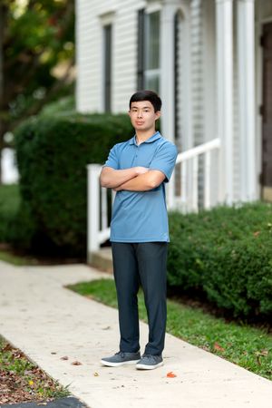Young man in blue polo standing confidently with arms crossed on suburban sidewalk.