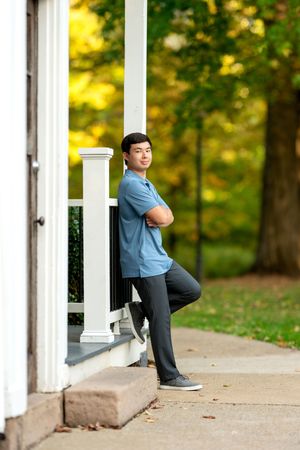 Young man in blue polo leaning against white porch column with autumn trees behind.