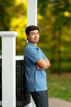 Young man with arms crossed leaning against white railing with green trees background.