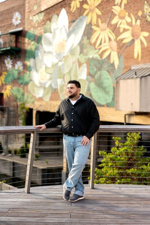 Young man in black shirt and jeans leaning on bridge railing near colorful floral mural.