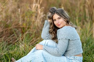 Senior portrait of smiling teen girl in gray sweater sitting in lush green grass field.