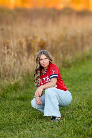 Teen girl in red varsity jersey crouching in green field with golden autumn background.