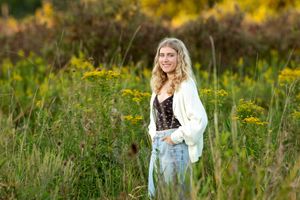 Smiling blonde girl in cream cardigan standing among wildflowers in autumn field.