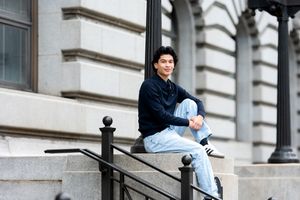 A young man in a dark hoodie and light jeans sits relaxed on stone steps outside a classic building.