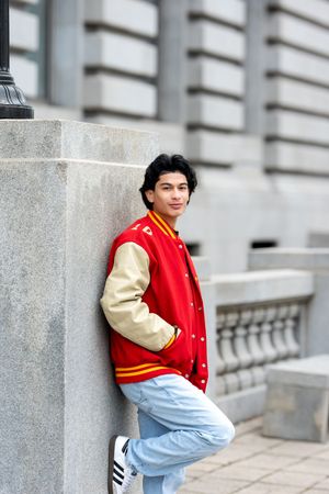 A teenage boy in a red varsity jacket and jeans leans against a stone column outside a grand building.