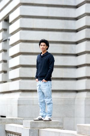 A young man in a navy polo and light jeans stands confidently on stone steps outside a historic building.