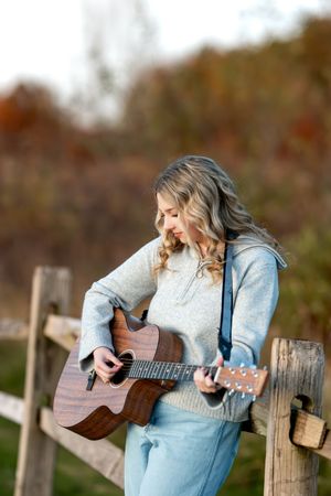 A teenage girl in a gray sweater plays an acoustic guitar while leaning on a wooden fence outdoors.