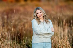 A smiling teenage girl in a gray sweater stands with arms crossed in a golden autumn field.