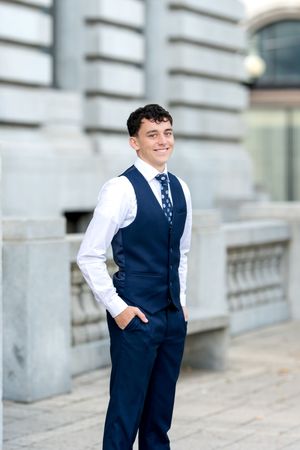Smiling young man in navy blue vest and tie posing outdoors near stone building.
