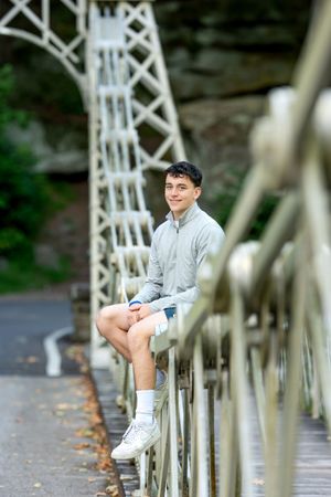 Young man in casual gray pullover sitting on white iron bridge railing outdoors.