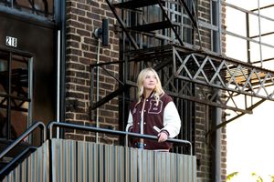 Blonde girl in varsity jacket standing on urban fire escape staircase of brick building.