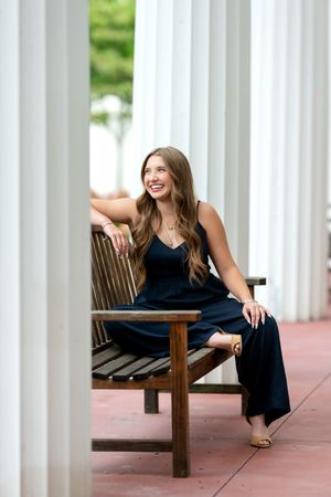 Laughing brunette teen girl in navy jumpsuit sitting on wooden bench between white columns.