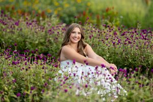 Smiling brunette teen girl in white dress sitting among colorful purple wildflowers for senior portrait.