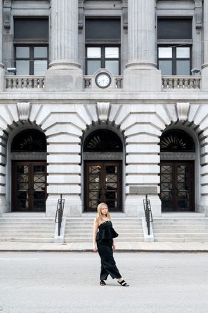 Blonde girl in black jumpsuit walks in front of grand neoclassical courthouse building exterior.