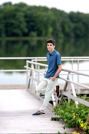 Teenage boy in blue polo shirt posing on a wooden lakeside dock with lush green trees in background.