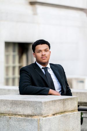 Young man in black suit and tie leaning on stone ledge outside classic building, serious expression.