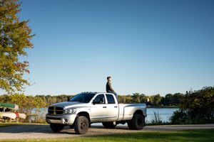 Person standing on top of silver pickup truck with lake and blue sky backdrop.