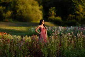 Elegant woman in a flowing red dress standing among tall wildflowers in a lush green garden at sunset.