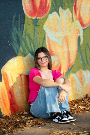 Teen girl with glasses in pink top sitting cross-legged against tulip mural wall outdoors.