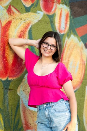 Smiling teen girl in pink top and glasses posing against colorful floral mural background.