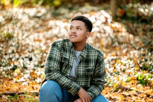 Young man in green plaid flannel shirt sitting among autumn leaves, gazing upward thoughtfully.