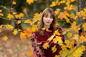 Young woman in burgundy dress surrounded by colorful fall foliage in an autumn forest setting.