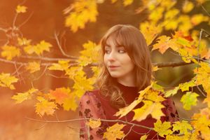 Young woman with bangs gazing at golden autumn leaves in a warm sunlit outdoor setting.