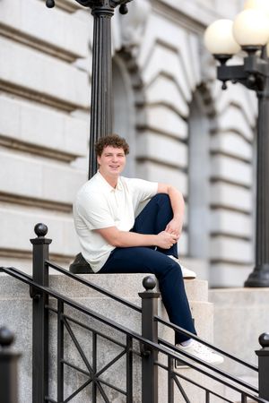 Smiling young man in white polo sitting on a stone railing beside an ornate lamppost.