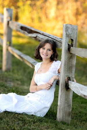 Young woman in white dress sitting on grass leaning against a weathered wooden fence outdoors.