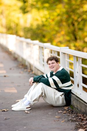 Smiling teen boy in green striped sweatshirt sitting against a white fence in autumn park.