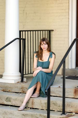 Young woman in a teal dress sitting gracefully on stone steps beside white columns and black railings.