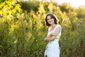 Smiling young woman in white dress standing among tall green wildflowers in warm sunlight.