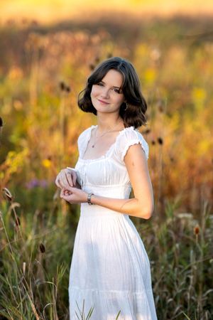 Young woman in white dress standing in a sunlit field of tall wildflowers at golden hour.