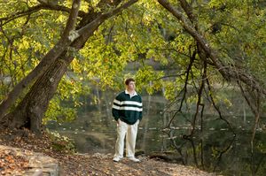 Young man in green striped shirt standing near a tree by a peaceful autumn lakeside.