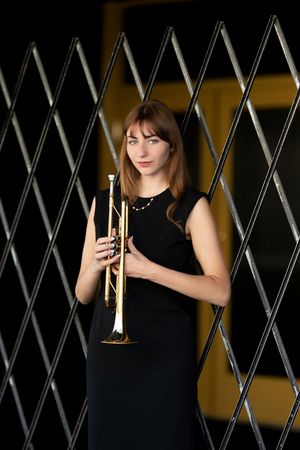 Young woman in black dress holding a gold trumpet, posing in front of a geometric metal gate.
