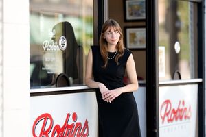Young woman in black dress leaning against the entrance of Robins restaurant on a sunny day.