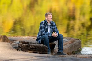 Young man in plaid shirt sits on rocks by a reflective lake with autumn foliage backdrop.
