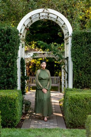 Woman in olive green dress stands under white garden arch surrounded by lush hedges and greenery.
